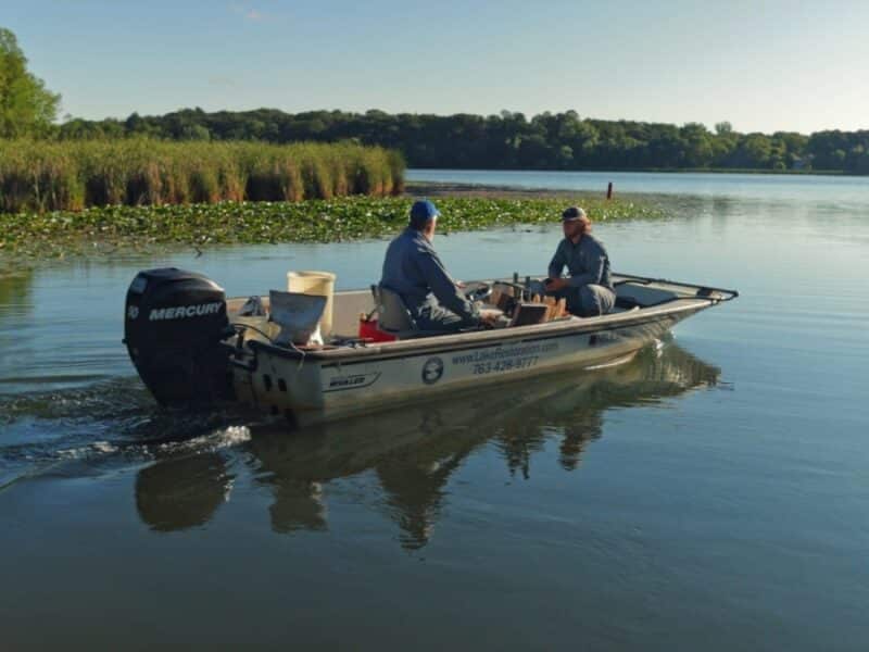 2 men in a Lake Restoration boat with shoreline weeds